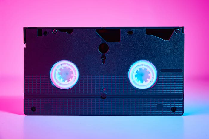 A vintage VHS tape with visible reels, set against a vibrant pink and blue gradient background.